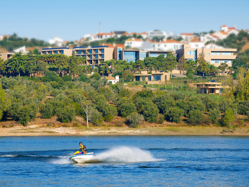 Ap Lago Montargil - Conference & Spa المظهر الخارجي الصورة The photo depicts a person riding a jet ski on a body of water. In the background, there are modern buildings and lush greenery, suggesting a scenic landscape, likely near a resort or upscale residential area. The clear blue sky and calm water enhanc