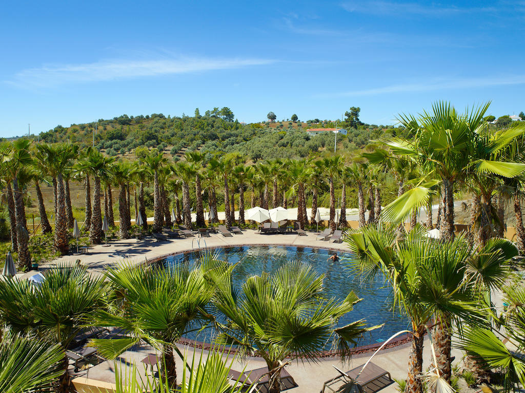 Ap Lago Montargil - Conference & Spa المظهر الخارجي الصورة The photo depicts a beautiful outdoor area featuring a circular swimming pool surrounded by palm trees. The pool area has several lounge chairs arranged around it. In the background, there is a lush green hillside, and the sky is clear with a bright 
