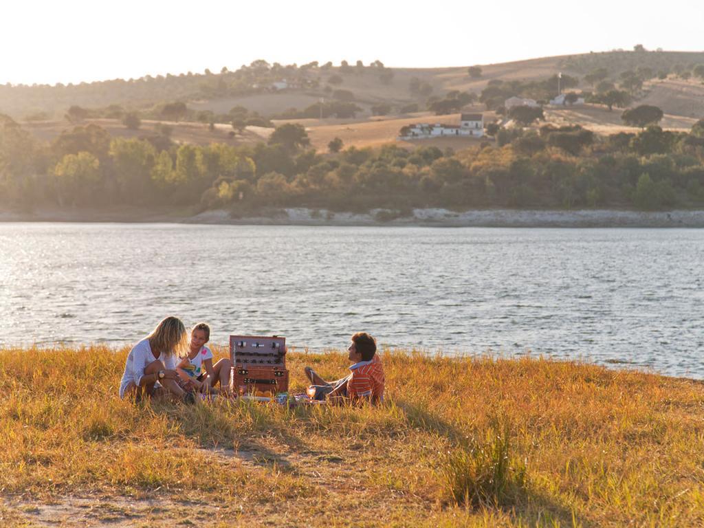 Ap Lago Montargil - Conference & Spa المظهر الخارجي الصورة The photo depicts a serene lakeside scene during sunset. Three people are gathered on a grassy area near the water, enjoying a picnic. They are seated around a picnic basket, with the shimmering lake in the background and hills in the distance. The w