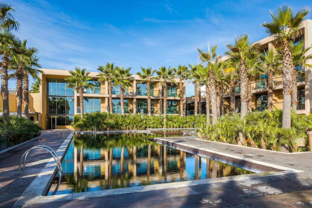 Ap Lago Montargil - Conference & Spa المظهر الخارجي الصورة The photo features a modern building surrounded by lush greenery and palm trees. In the foreground, there is a serene swimming pool with a reflective surface that captures the surrounding environment. The architecture of the building appears to have 