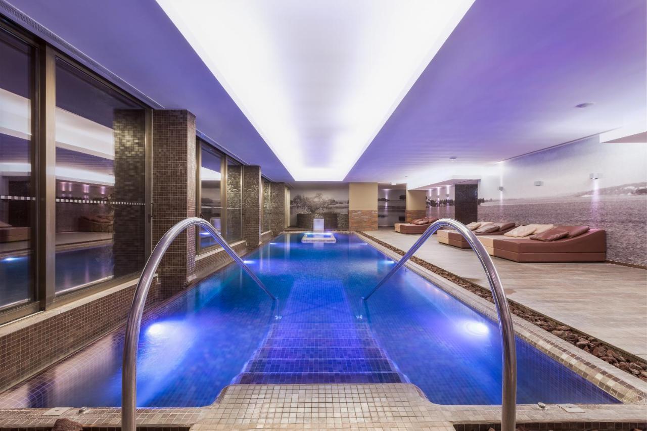 Ap Lago Montargil - Conference & Spa المظهر الخارجي الصورة The photo shows a modern indoor swimming pool area. The pool features a sleek design with steps leading into the water and a gentle curve at the sides. The water is illuminated in a blue hue, enhancing the relaxing atmosphere. Surrounding the pool ar