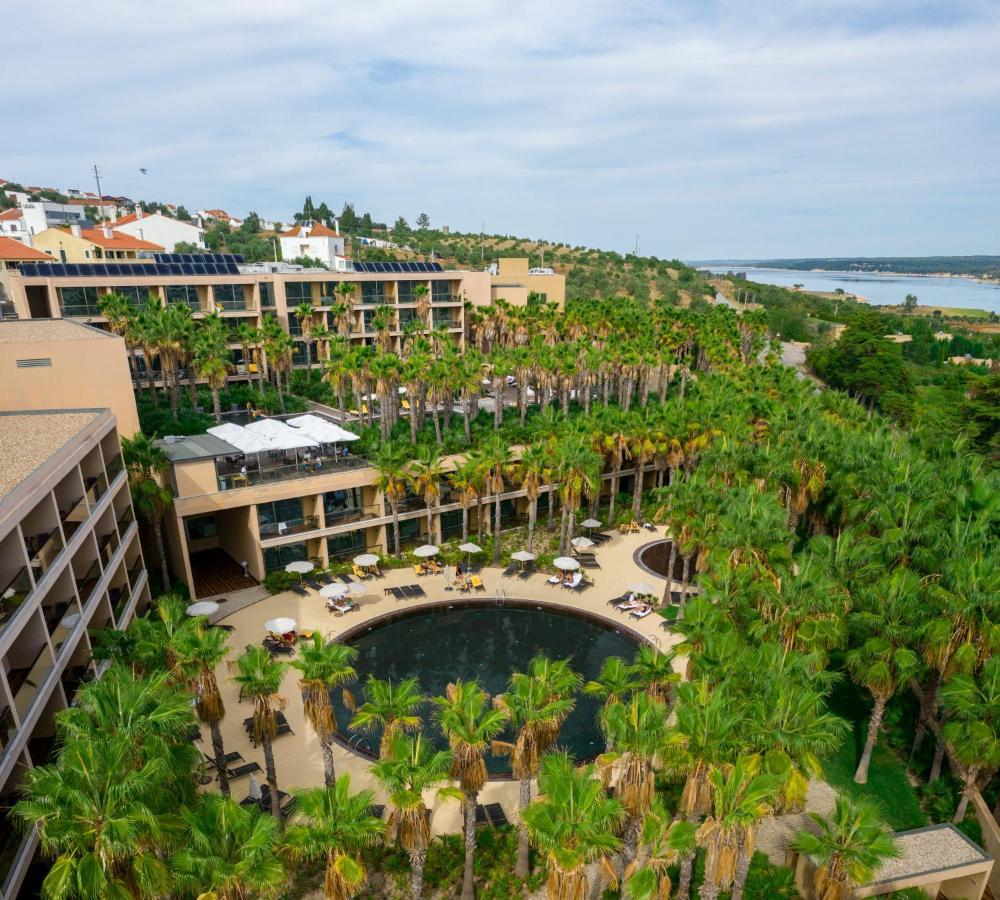 Ap Lago Montargil - Conference & Spa المظهر الخارجي الصورة The photo shows a modern hotel complex set amidst lush greenery, featuring numerous palm trees. In the foreground, there is a circular swimming pool surrounded by lounge chairs. The hotel itself has multiple levels with balconies overlooking the pool
