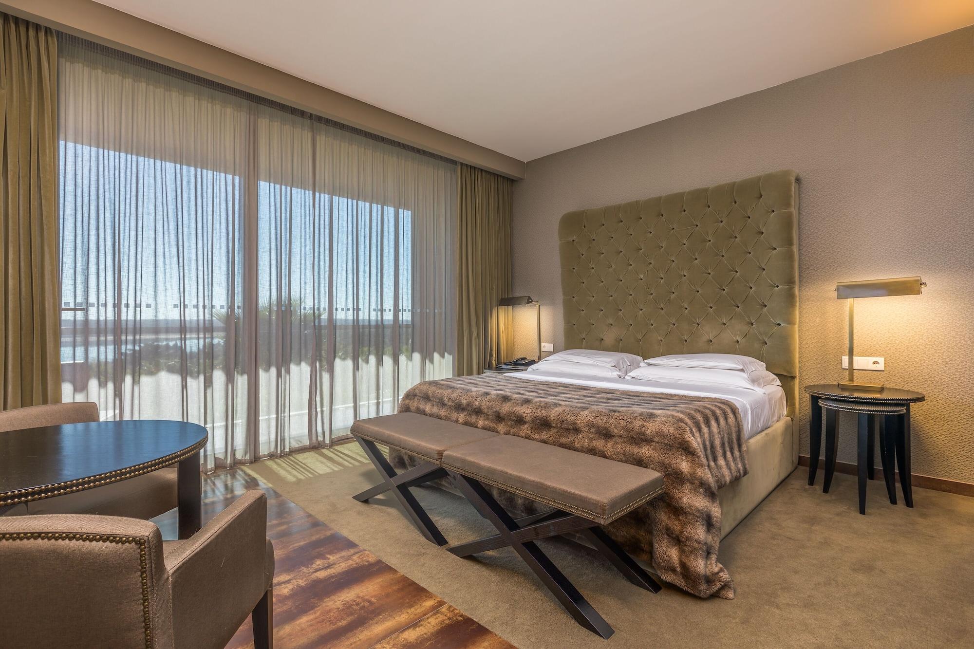 Ap Lago Montargil - Conference & Spa المظهر الخارجي الصورة The photo shows a modern hotel room featuring a large bed with a tufted headboard and a cozy blanket. There are curtains draped over large windows that allow natural light to flow in, providing a view of the outside. A small seating area includes two