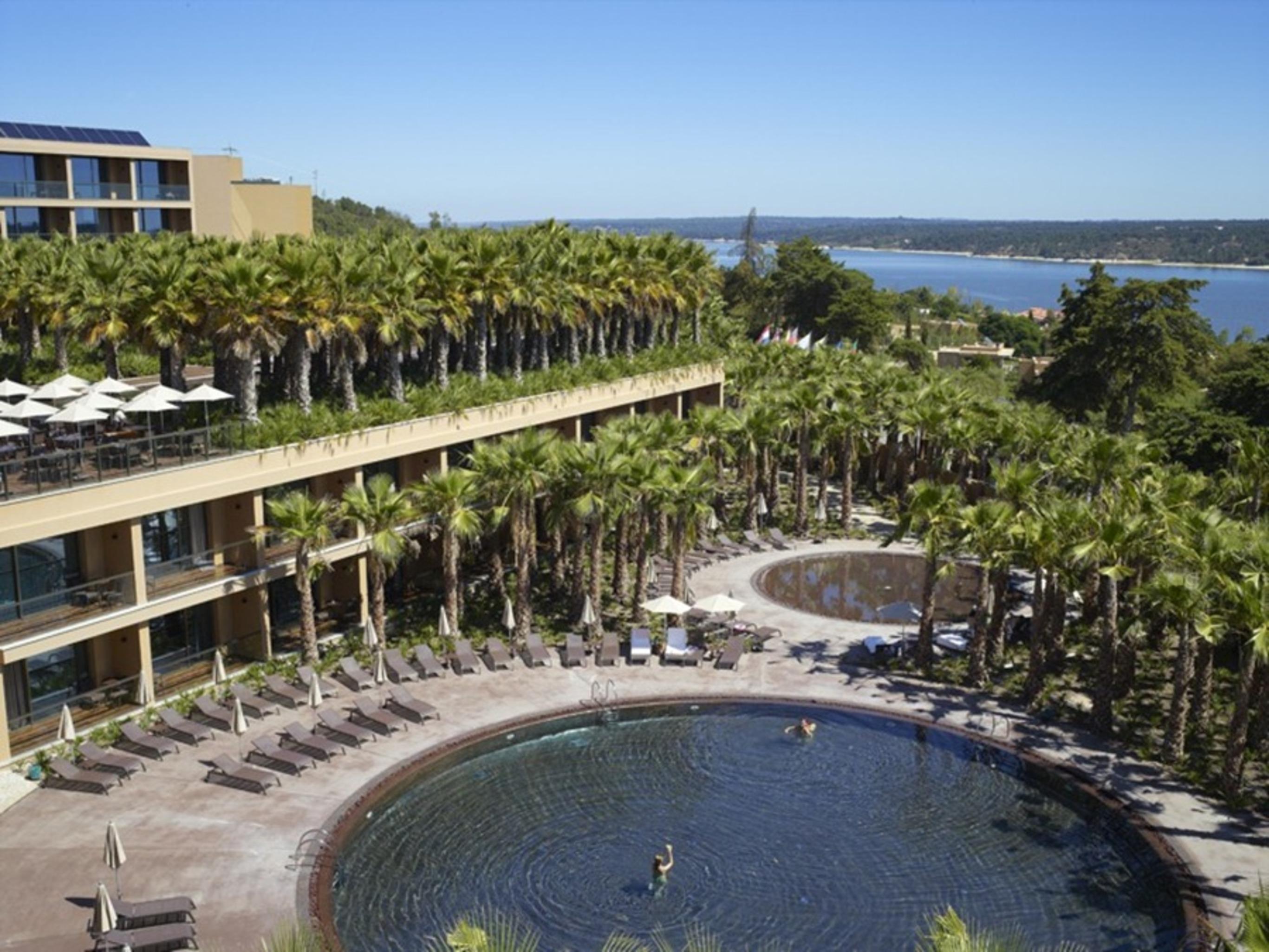 Ap Lago Montargil - Conference & Spa المظهر الخارجي الصورة The photo shows a scenic view of a resort area. In the foreground, there are circular pools surrounded by lounge chairs, with a few people enjoying the water. Palm trees line the edges, creating a tropical atmosphere. In the background, there is a mu