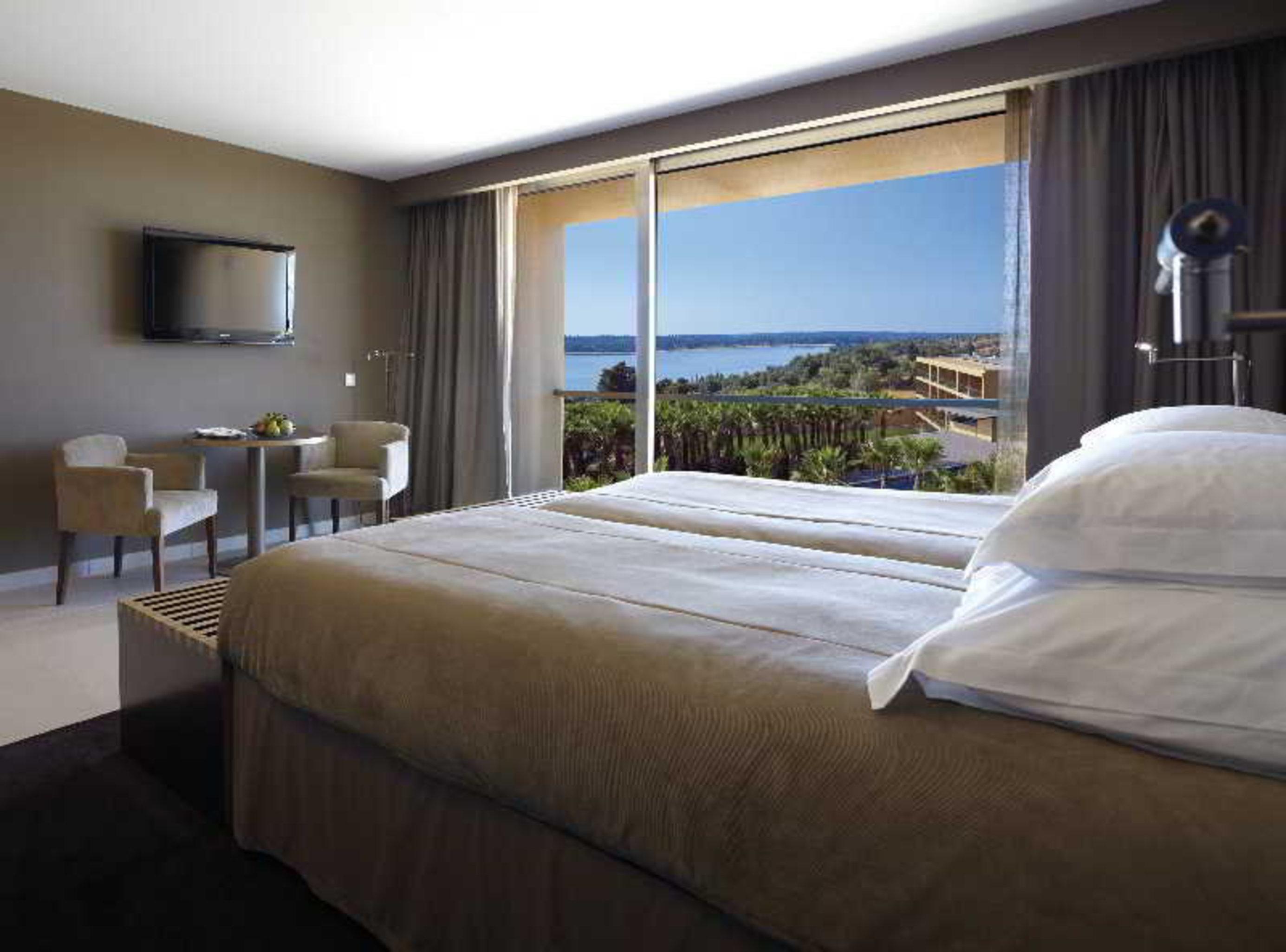 Ap Lago Montargil - Conference & Spa المظهر الخارجي الصورة The photo shows a modern hotel room with a large window offering a view of the sea and mountains. The room features a comfortable bed with white pillows and brown bedding. There is a small sitting area with two chairs and a table, as well as a wall-m
