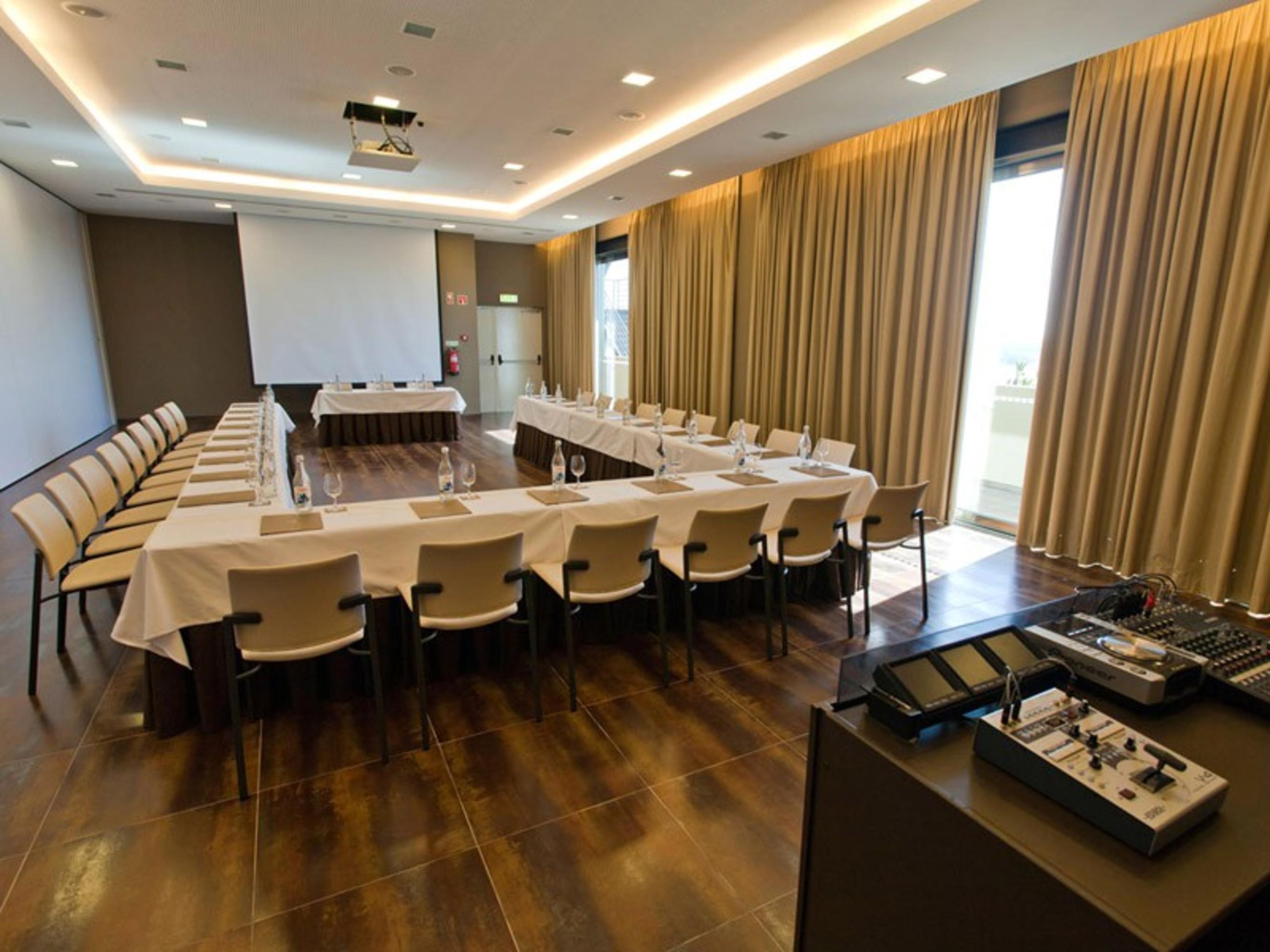 Ap Lago Montargil - Conference & Spa المظهر الخارجي الصورة The photo shows a modern conference room set up for a meeting. There are several tables arranged in a U-shape, surrounded by chairs. A large screen and projector are positioned at the front, with a table for speakers or presenters. The room has large