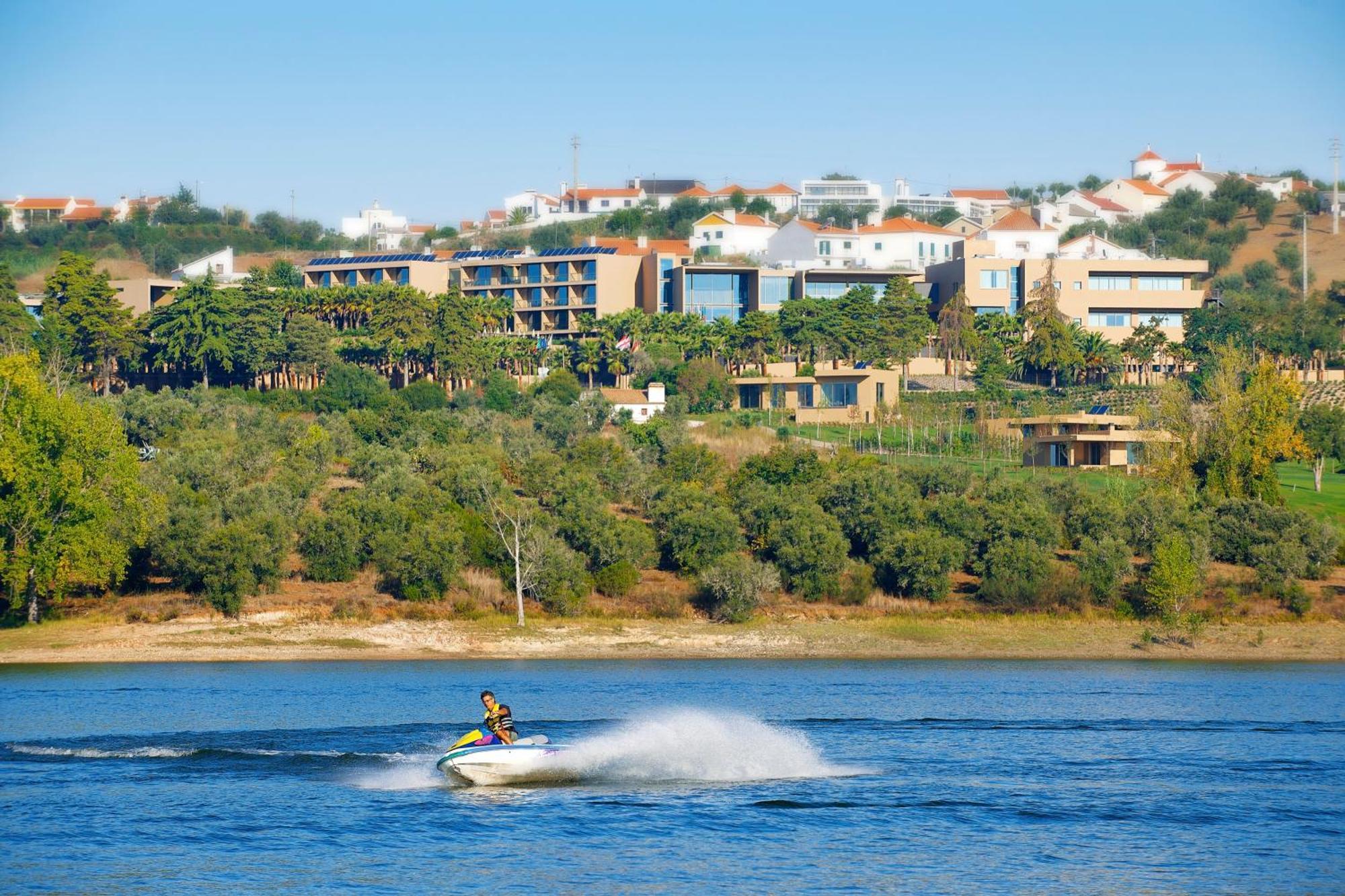 Ap Lago Montargil - Conference & Spa المظهر الخارجي الصورة The photo shows a person riding a jet ski on a body of water, creating a spray as they speed across the surface. In the background, there are modern buildings and a hillside with trees and greenery. The sky is clear and blue, suggesting a bright and 