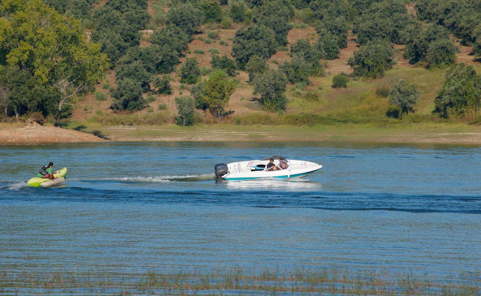 Ap Lago Montargil - Conference & Spa المظهر الخارجي الصورة The photo shows a scene on a water body, likely a river or lake. In the foreground, a speedboat is moving quickly through the water, leaving a wake behind it. There are two people visible in the boat. In the background, there is a hilly area covered 