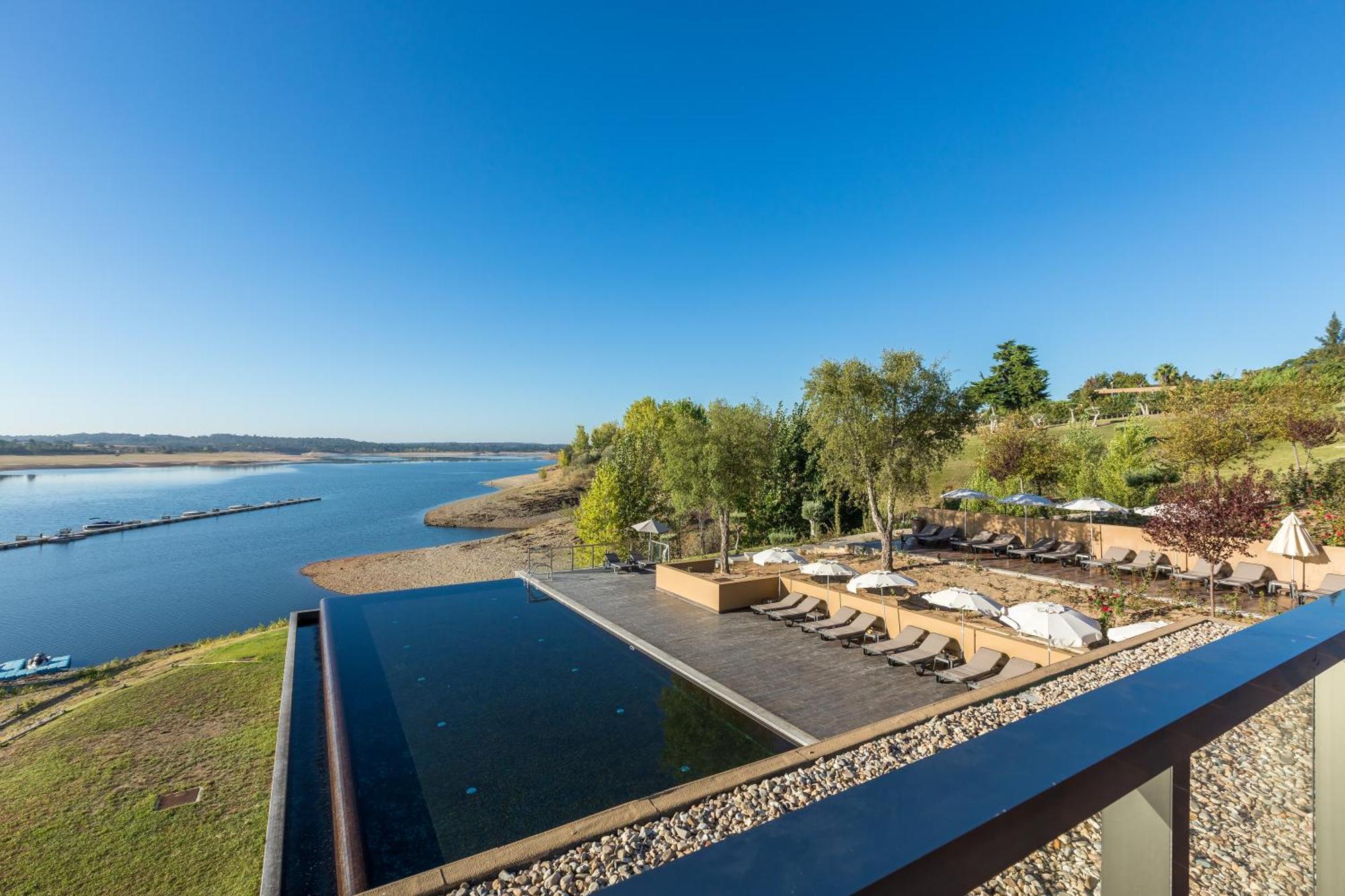 Ap Lago Montargil - Conference & Spa المظهر الخارجي الصورة The photograph depicts a scenic view of a tranquil water body, likely a lake or river, bordered by lush greenery. In the foreground, there is an infinity pool that blends seamlessly with the water beyond. Comfortable lounge chairs are arranged on a w