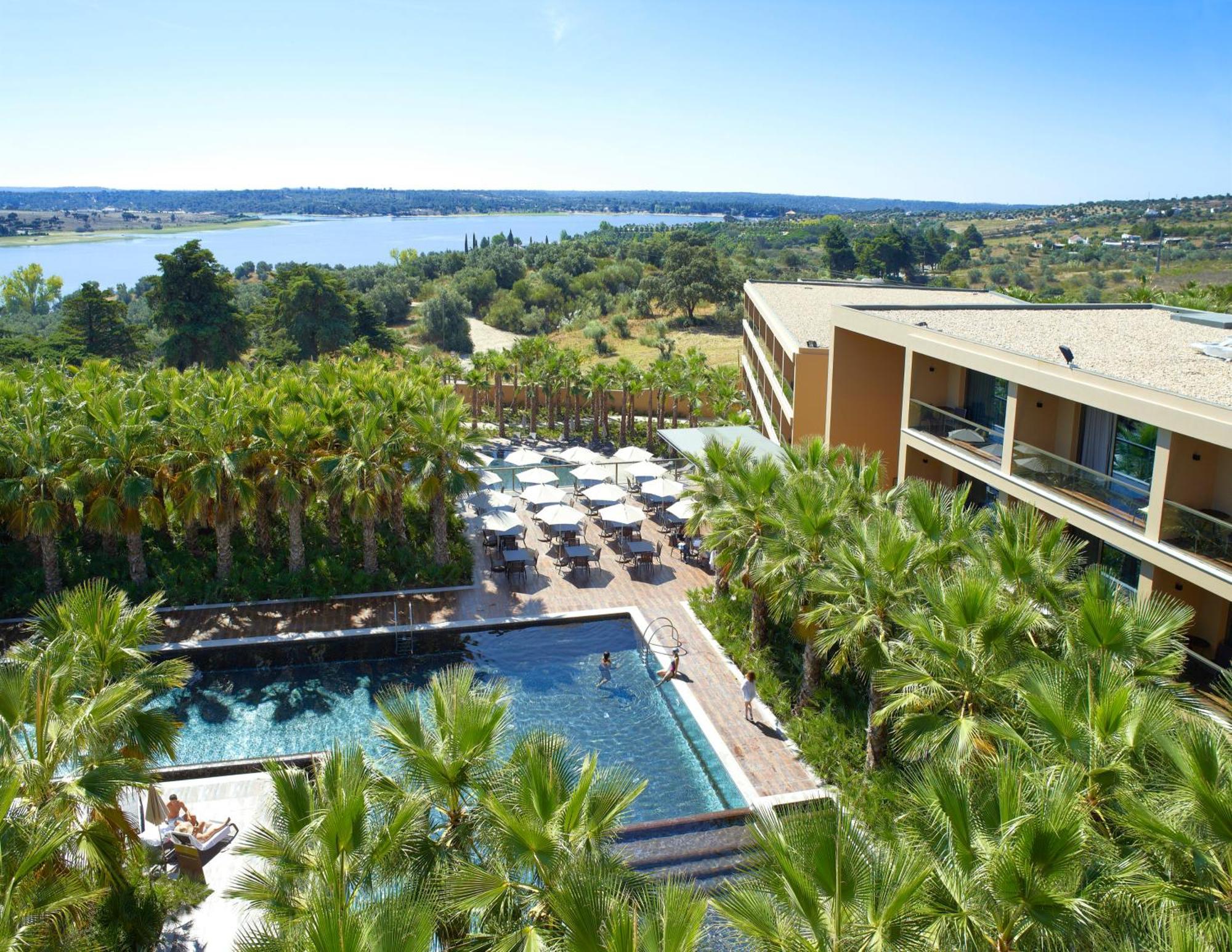 Ap Lago Montargil - Conference & Spa المظهر الخارجي الصورة The photo shows a scenic view of a modern hotel or resort. In the foreground, there is a swimming pool surrounded by palm trees, with lounge chairs positioned beside it. Sun umbrellas provide shade over the chairs. The background features a lush land
